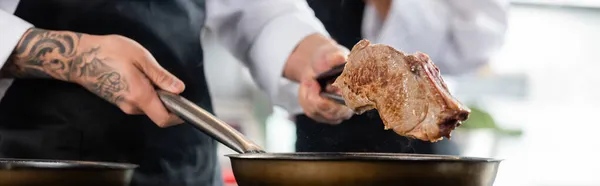 Cropped view of chef with tongs holding meat and frying pan in kitchen, banner — Stock Photo