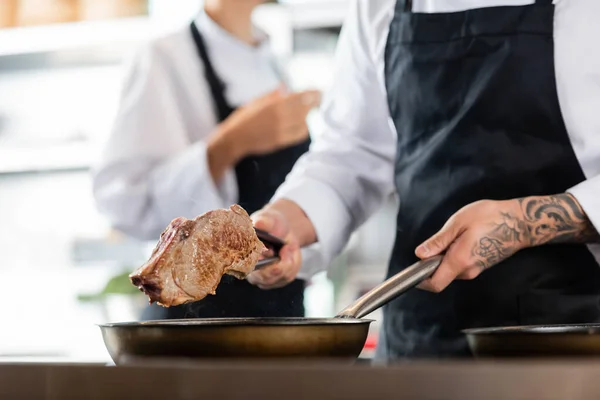 Vista recortada del chef en delantal sosteniendo carne cerca de la sartén en la cocina — Stock Photo