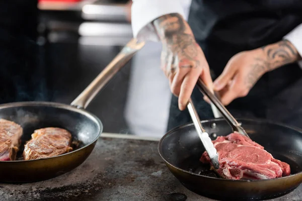 Cropped view of blurred chef with tongs roasting meat on cooktop in kitchen — Stock Photo