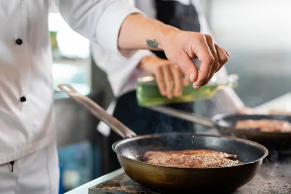 Vista cortada do chef derramando sal enquanto cozinha carne na frigideira na cozinha — Fotografia de Stock