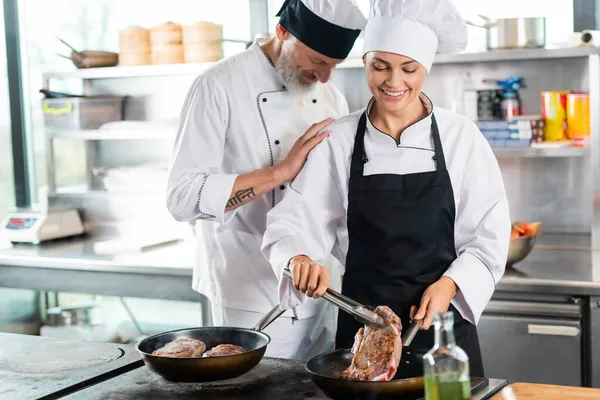 Chef debout près d'un collègue souriant rôtissant la viande dans la cuisine — Stock Photo