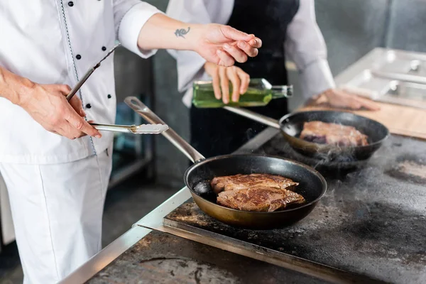 Обрізаний вид шеф-кухаря, який тримає щипці під час обсмажування м'яса на кухонній плиті — стокове фото