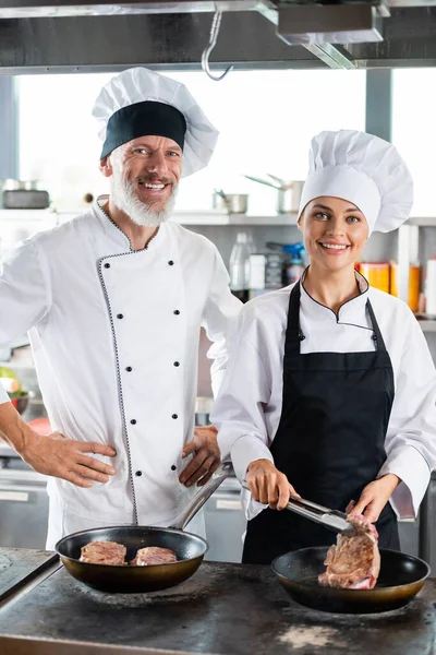 Chefs sonrientes en uniforme mirando a la cámara mientras cocinan carne en la cocina - foto de stock