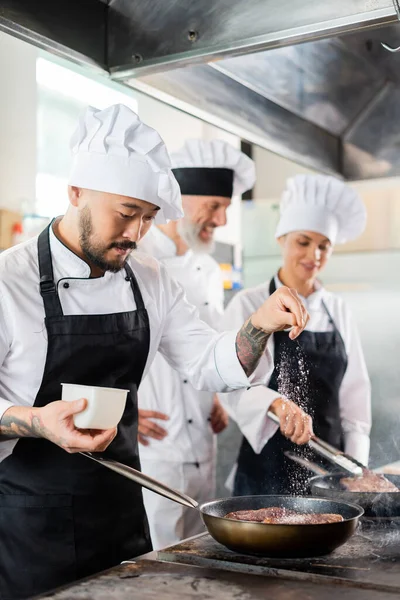 Chef asiático derramando sal na carne no fogão perto de colegas embaçados na cozinha — Fotografia de Stock