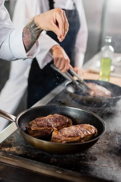 Cropped view of tattooed chef pouring salt while roasting meat on cooktop in kitchen — Stock Photo