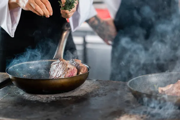 Vue recadrée du chef versant du romarin tout en rôtissant la viande dans la cuisine — Photo de stock