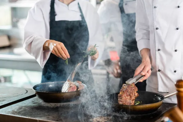 Cropped view of chefs with tongs and rosemary roasting meat in kitchen — Stock Photo