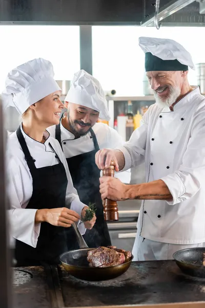 Улыбающийся шеф-повар приправляет мясо на сковородке рядом с веселыми многонациональными коллегами с розмарином на кухне — стоковое фото
