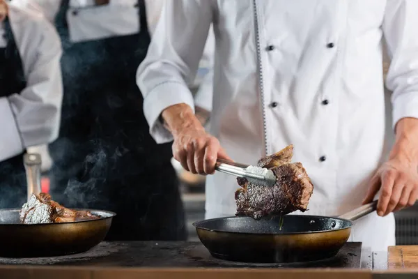 Cropped view of chef holding meat in tongs near frying pans and colleagues in kitchen — Stock Photo