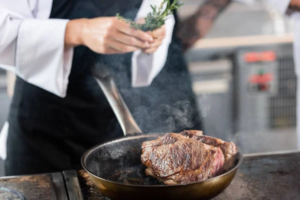 Roasted meat on frying pan near blurred chef holding rosemary in kitchen — Stock Photo