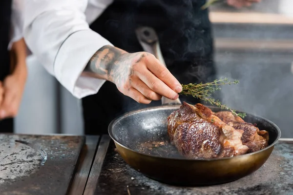 Cropped view of tattooed chef holding rosemary near roasted meat on frying pan — Stock Photo