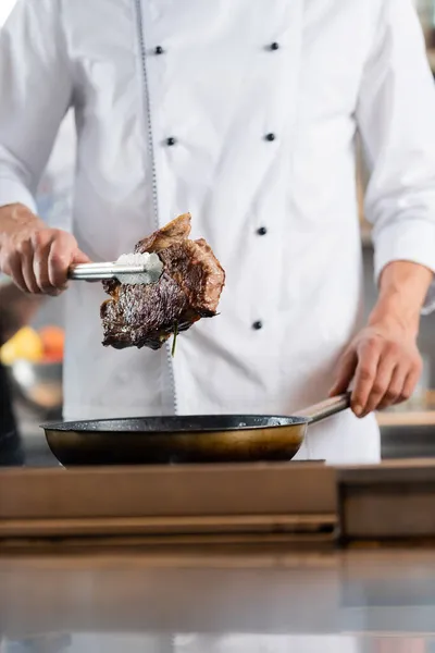 Vue recadrée du chef tenant de la viande rôtie dans des pinces près de la poêle — Photo de stock