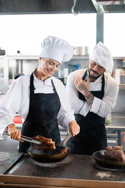 Chef asiático de pie cerca de compañero de cocina de carne en la cocina - foto de stock