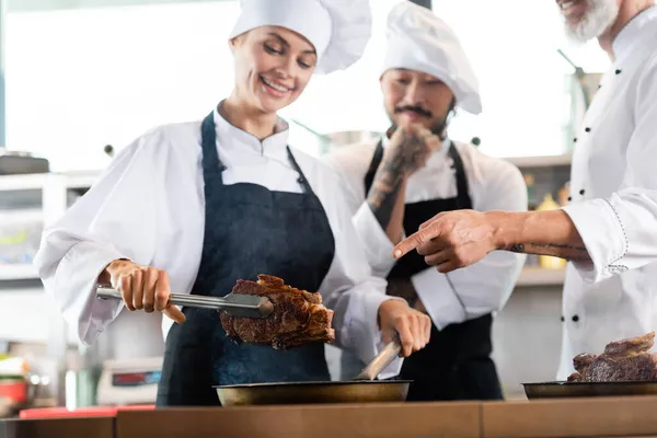 Chef pointing at roasted steak near multiethnic colleagues in aprons in kitchen — Stock Photo