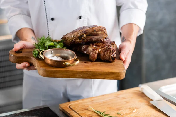 Cropped view of chef holding roasted meat on cutting board in kitchen — Stock Photo