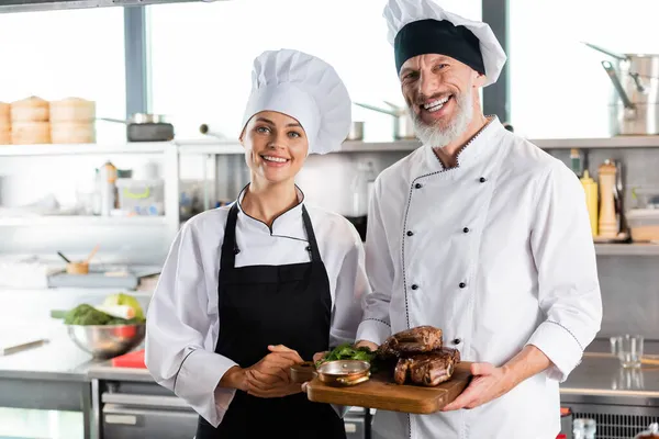 Positive chefs smiling at camera while holding roasted meat in kitchen — Stock Photo