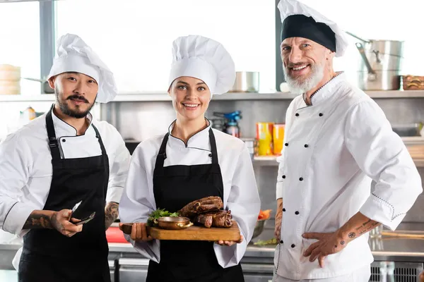 Усміхнений шеф-кухар тримає смажене м'ясо на обробній дошці біля міжрасових колег на кухні — стокове фото