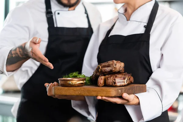 Обрезанный вид на повара, указывающий на вкусное мясо на разделочной доске рядом с коллегой на кухне — стоковое фото