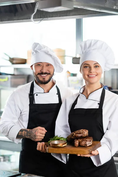 Tattooed asian chef standing near colleague with roasted meat on cutting board in kitchen — Stock Photo