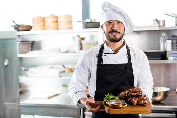 Улыбающийся азиатский шеф-повар держит разделочную доску с жареным мясом и зеленью на кухне — стоковое фото
