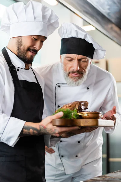 Mature chef smelling roasted meat near asian colleague in kitchen — Stock Photo