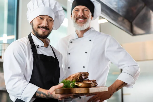 Sorrindo asiático chef segurando placa de corte com carne assada perto colega na cozinha — Fotografia de Stock