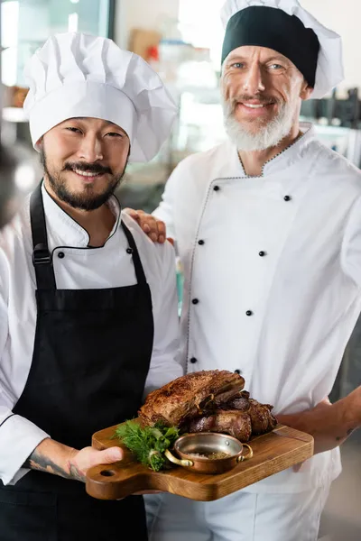 Chefs multiétnicos positivos sosteniendo carne cocida en la tabla de cortar en la cocina - foto de stock