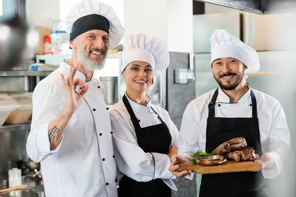 Chef sorrindo mostrando gesto ok perto de colegas inter-raciais com carne assada na cozinha — Fotografia de Stock