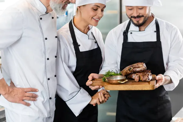 Sorrindo chefs inter-raciais olhando para carne saborosa em tábua de corte na cozinha — Fotografia de Stock