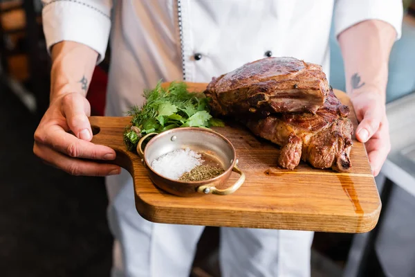 Cropped view of chef in uniform holding cutting board with spices and roasted meat — Stock Photo