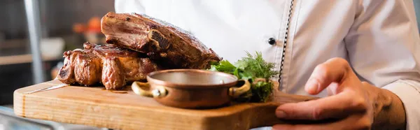 Cropped view of chef holding cutting board with roasted meat and greens in kitchen, banner — Stock Photo