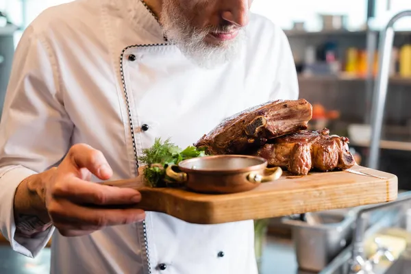 Vue recadrée du chef tenant la planche à découper avec steaks et légumes verts dans la cuisine — Photo de stock