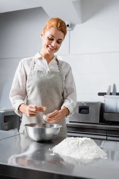 Smiling chef holding egg near flour and bowl in kitchen — Stock Photo
