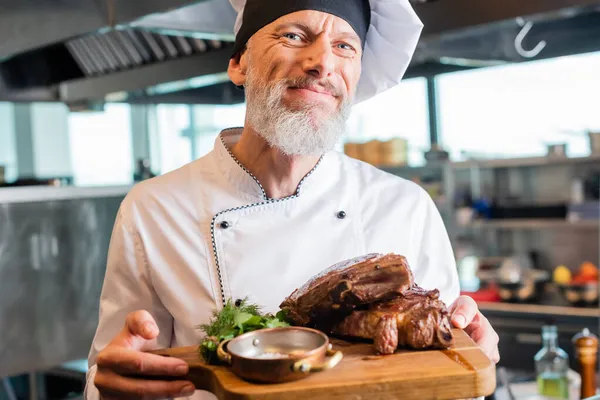 Reifer Koch lächelt in die Kamera, während er in der Küche gebratenes Fleisch auf dem Schneidebrett hält — Stockfoto