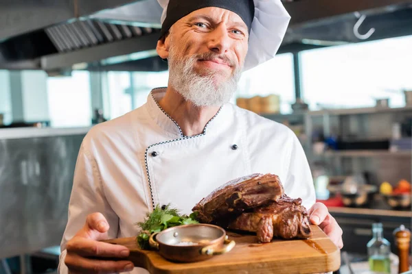 Chef mature souriant à la caméra tout en tenant de la viande rôtie sur la planche à découper dans la cuisine — Photo de stock