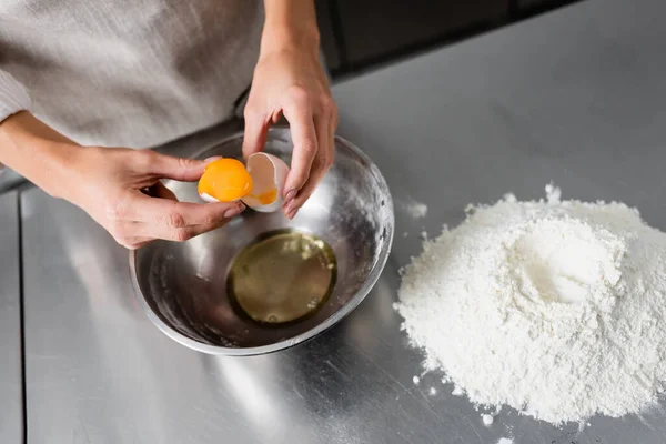 Обрізаний вид шеф-кухаря, що тримає яйце та яєчну шкаралупу біля миски та борошна на столі — стокове фото
