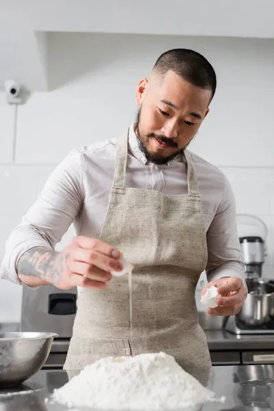 Asiatischer Koch in Schürze gießt in Küche Ei auf Mehl — Stockfoto