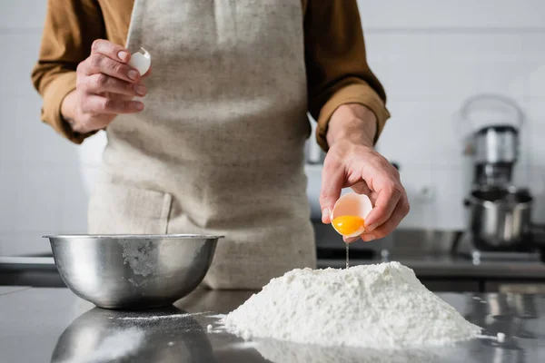 Vue recadrée du chef dans le tablier verser l'oeuf sur la farine près du bol sur la table — Photo de stock