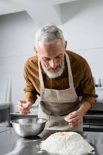 Mature chef holding eggshell near flour and bowl on table — Stock Photo