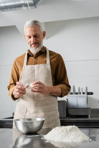 Lächelnder älterer Koch mit Eierschale in der Nähe von Mehl und Schüssel auf dem Tisch, während er in der Küche kocht — Stockfoto