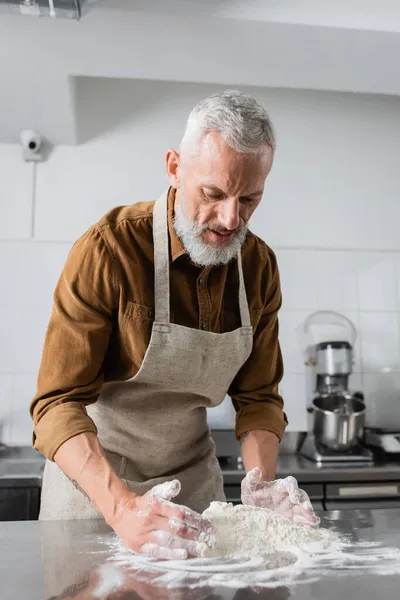 Mature bearded chef in apron making dough on kitchen table — Stock Photo