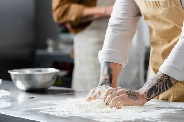 Cropped view of tattooed chef making dough near blurred colleague in kitchen — Stock Photo