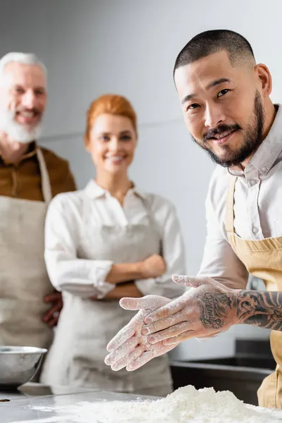 Asian chef smiling near flour and blurred colleagues in kitchen — Stock Photo