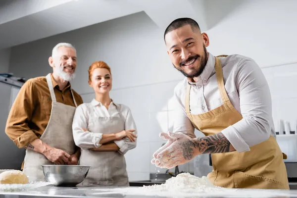 Позитивний азіатський шеф-кухар в фартусі, що стоїть біля борошна і розмитих колег на кухні — стокове фото