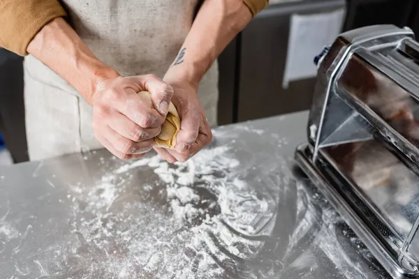 Vista recortada del chef sosteniendo masa cerca de la máquina de hacer pasta y harina en la mesa - foto de stock