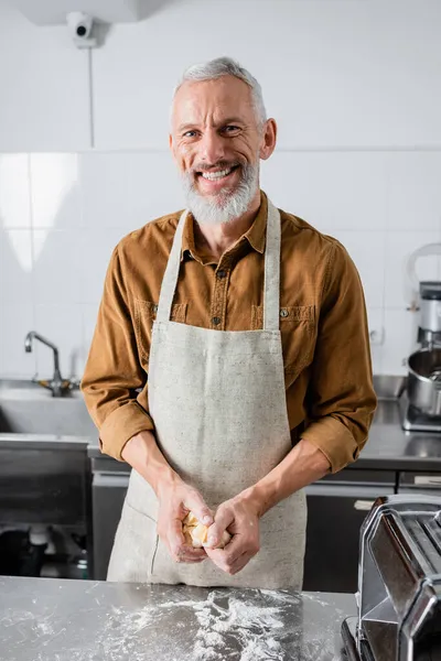 Chef souriant dans le tablier tenant la pâte et regardant la caméra près de la machine à pâtes dans la cuisine — Photo de stock