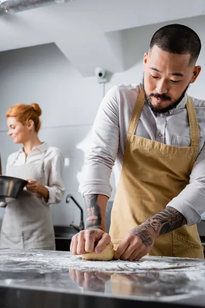 Chef asiático haciendo masa cerca borrosa colega con tazón en la cocina - foto de stock