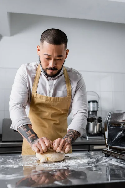 Татуированный азиатский шеф-повар в фартуке делает тесто возле машины для приготовления макарон на столе на кухне — стоковое фото