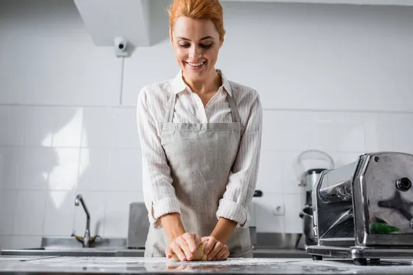 Chef sonriente en delantal haciendo masa cerca de la máquina de hacer pasta en la cocina del restaurante - foto de stock
