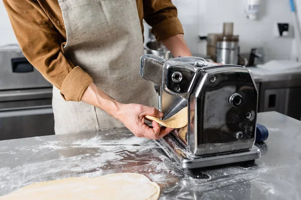 Vista ritagliata dello chef in grembiule con impasto vicino alla macchina per la pasta e farina in cucina — Stock Photo